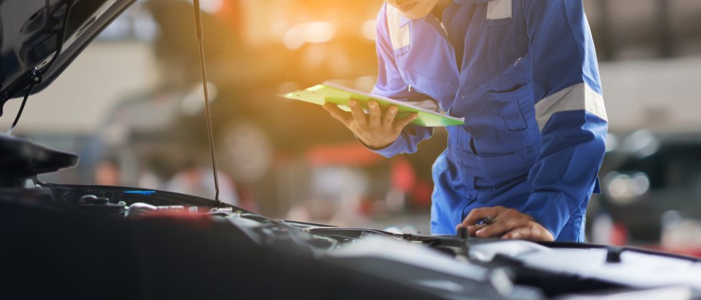 A mechanic in a blue uniform inspecting the engine bay of a vehicle, holding a clipboard and reviewing notes in an auto repair shop.